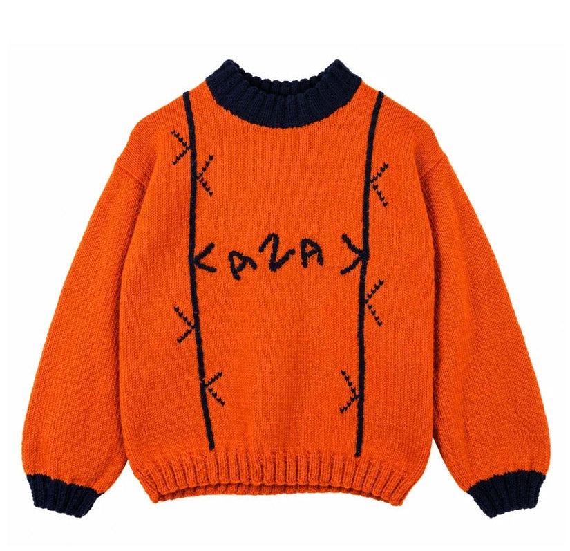 Bright orange knit sweater with dark navy blue ribbed collar, cuffs, and hem, featuring decorative black stitched details and the word 'KAZAK' knitted across the chest.
