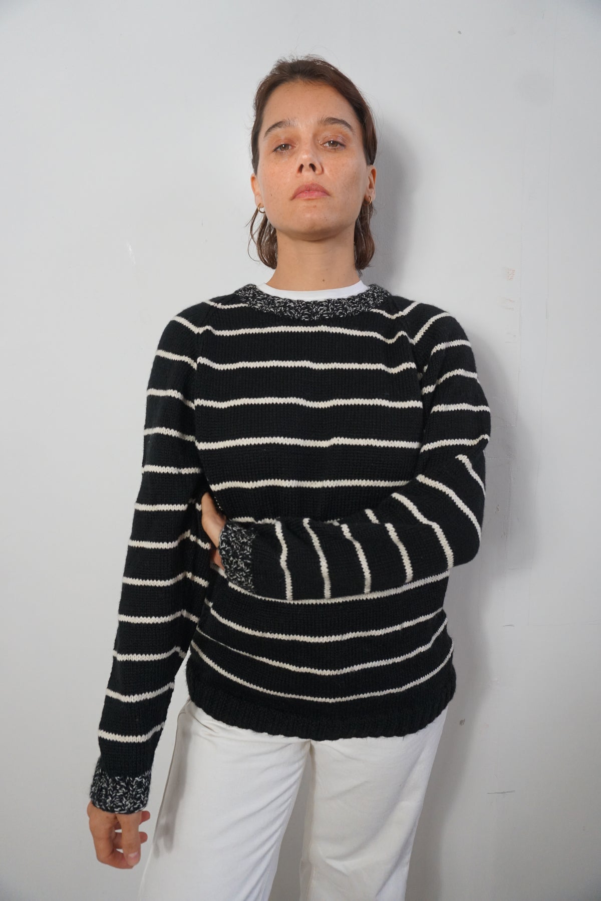 A woman wearing a black and white horizontally striped knit sweater with textured gray trim at the collar, cuffs, and pocket, paired with white pants, standing against a plain light gray wall.