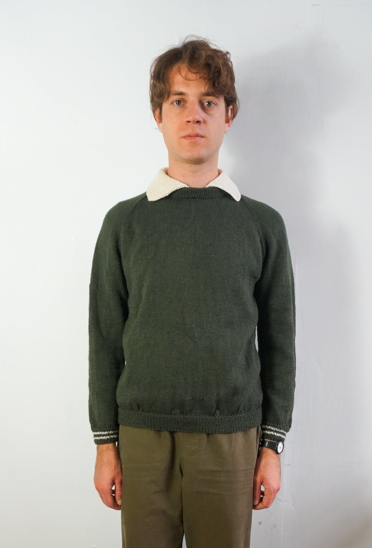 A man wearing a dark green knit sweater with a contrasting white collar and striped cuffs, paired with olive green trousers, standing against a plain white wall.