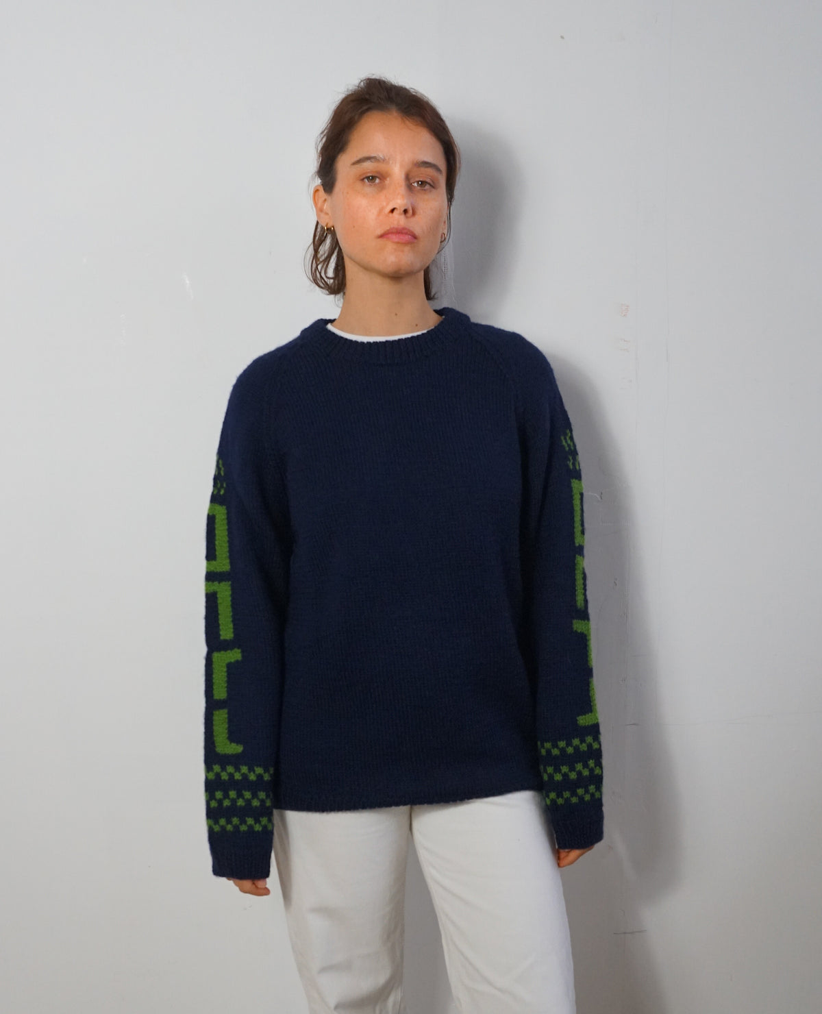 A woman wearing a navy blue knit sweater with green geometric patterns on the sleeves, paired with white pants, standing against a plain light gray wall.
