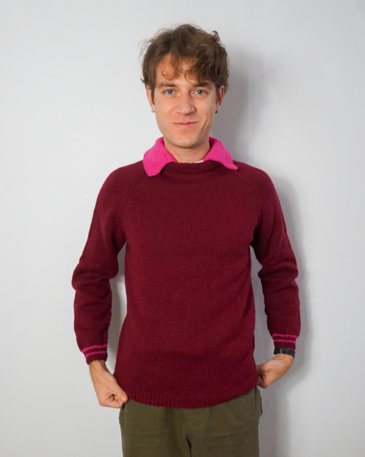A man wearing a burgundy knit sweater with a bright pink collar and matching pink ribbed cuffs, paired with olive green pants, standing against a light gray wall.