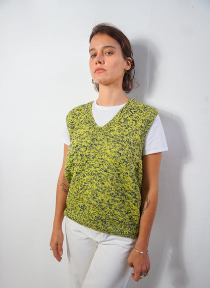 A woman wearing a lime green and black speckled knit sweater vest over a white t-shirt and white pants, standing against a plain white wall.