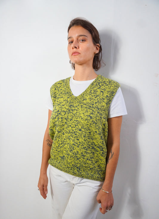 A woman wearing a lime green and black speckled knit sweater vest over a white t-shirt and white pants, standing against a plain white wall.