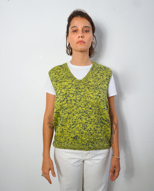 A woman modeling a lime green and black speckled knit sweater vest over a white t-shirt and white pants, standing against a plain light gray wall.