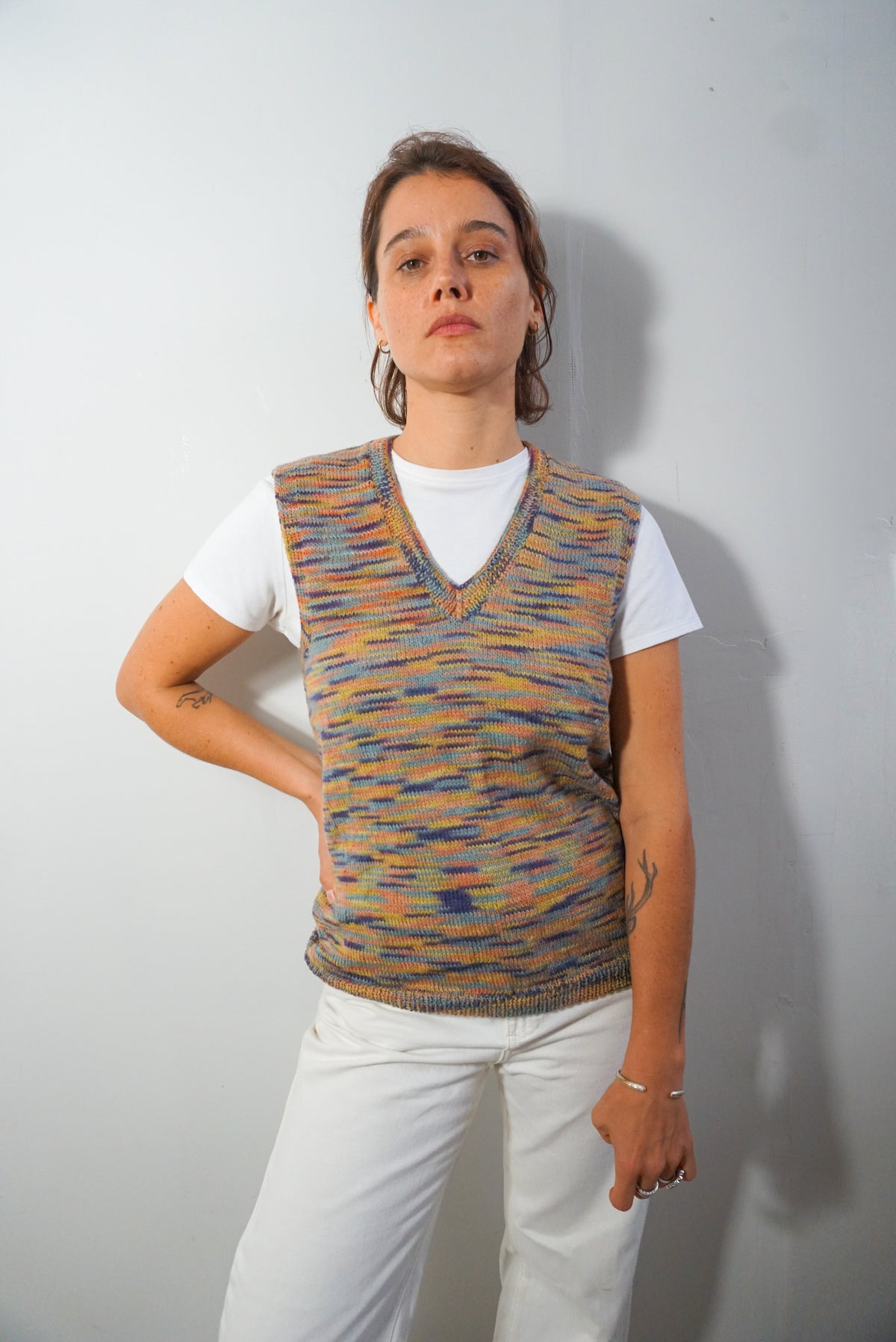 A woman wearing a multicolor knit sweater vest over a white t-shirt and white pants, standing against a plain light gray wall.