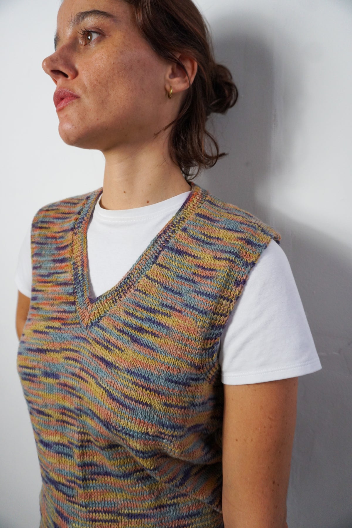 A woman wearing a sleeveless, V-neck knit vest with a multicolor striped pattern over a white t-shirt, photographed against a plain light gray wall.