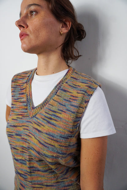 A woman wearing a sleeveless, V-neck knit vest with a multicolor striped pattern over a white t-shirt, photographed against a plain light gray wall.