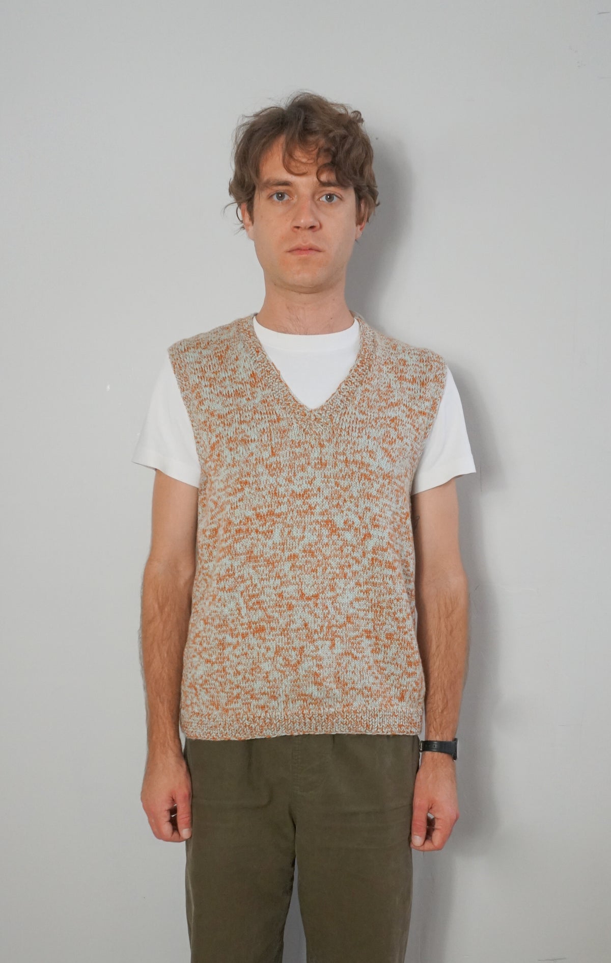A man wearing a sleeveless knit sweater vest with a speckled beige and orange pattern over a white t-shirt and olive green pants, standing against a plain light gray wall.