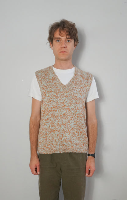 A man wearing a sleeveless knit sweater vest with a speckled beige and orange pattern over a white t-shirt and olive green pants, standing against a plain light gray wall.