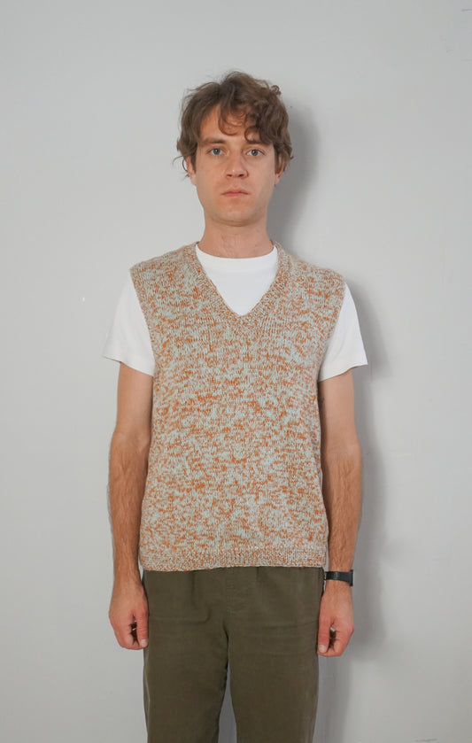 A man wearing a sleeveless knit sweater vest with a speckled beige and orange pattern over a white t-shirt and olive green pants, standing against a plain light gray wall.