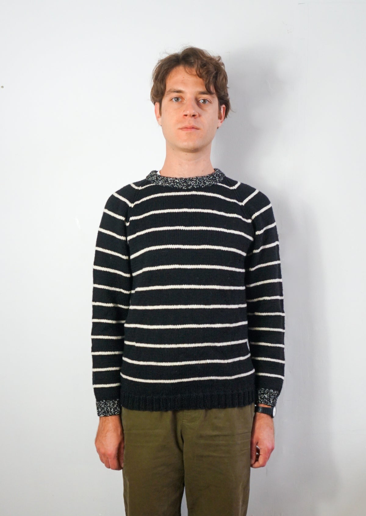 A man wearing a navy blue and white horizontally striped knit sweater with patterned trim at the collar, cuffs, and hem, paired with olive green trousers, standing against a plain white wall.
