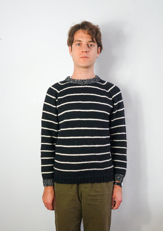 A man wearing a navy blue and white horizontally striped knit sweater with patterned trim at the collar, cuffs, and hem, paired with olive green trousers, standing against a plain white wall.