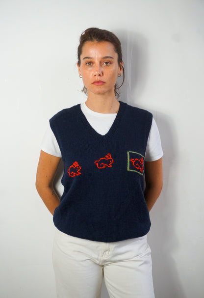 A woman wearing a navy blue knit sweater vest with red embroidered motifs and a green-bordered patch on the front, layered over a white t-shirt and white pants, standing against a plain light gray wall.