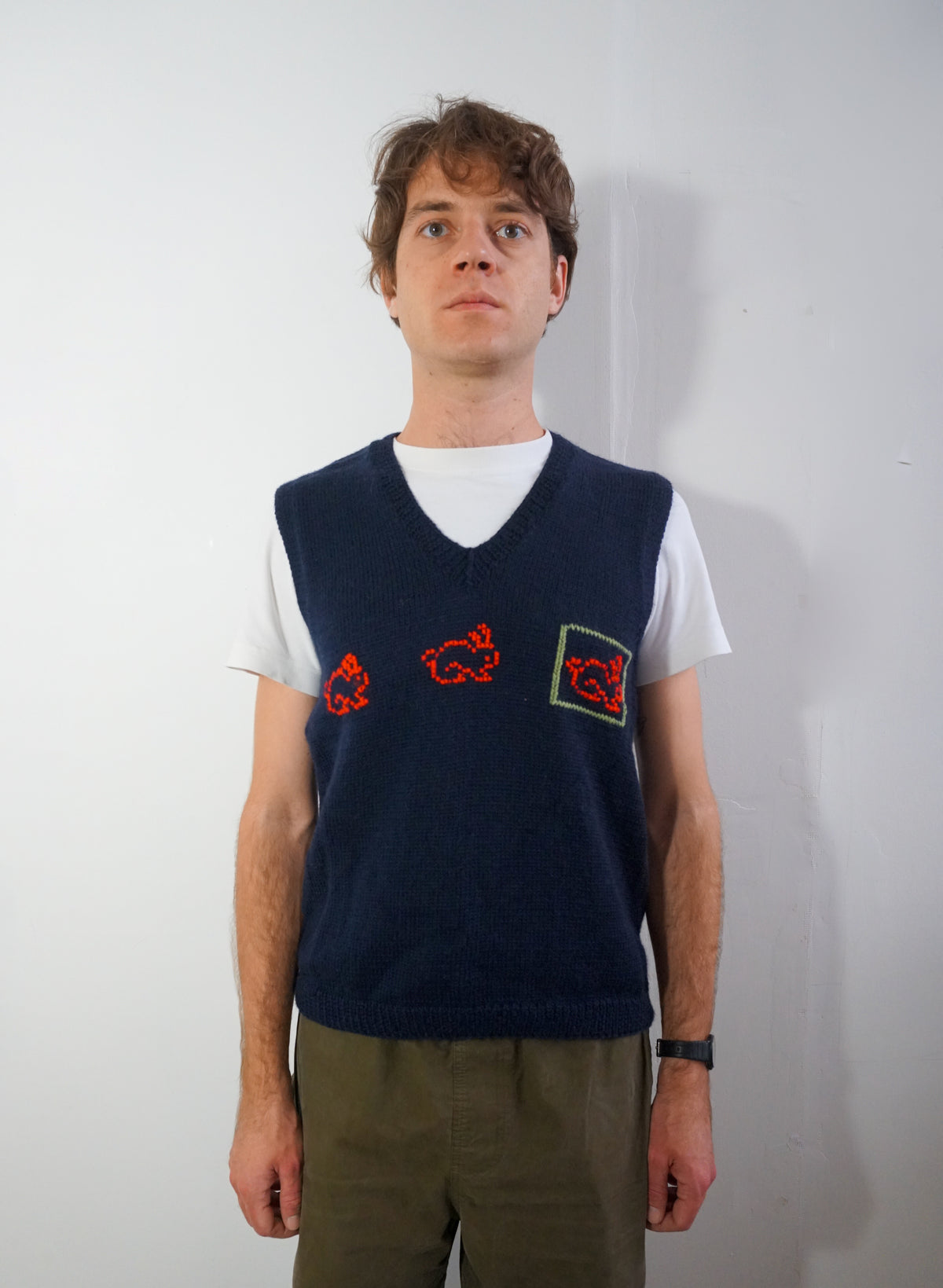 A man wearing a navy blue knit sweater vest over a white t-shirt and olive green pants, standing against a plain white wall.