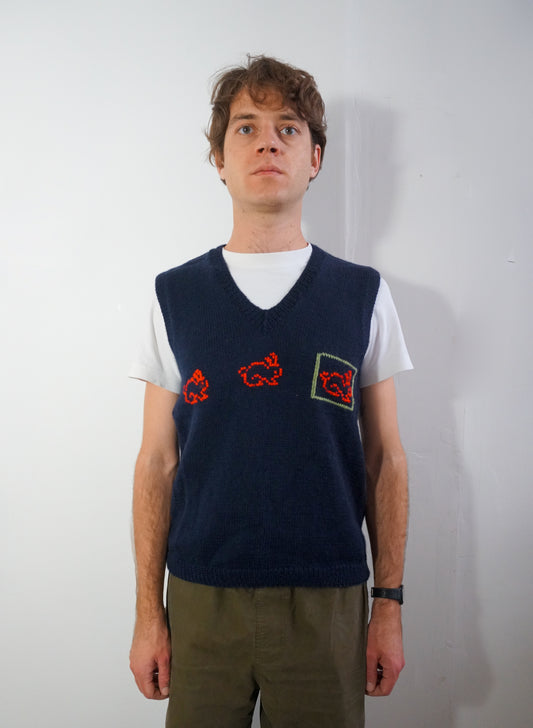 A man wearing a navy blue knit sweater vest over a white t-shirt and olive green pants, standing against a plain white wall.
