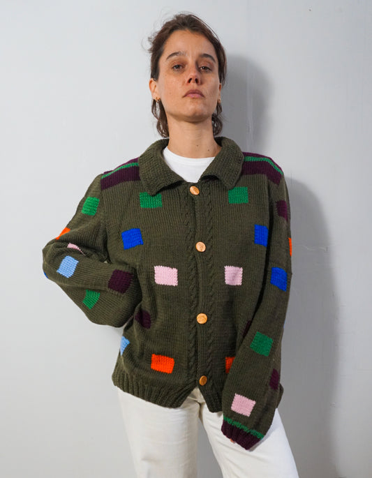 A woman wearing a dark olive green cable-knit cardigan with colorful geometric patchwork squares in blue, green, pink, orange, purple, and white, paired with white pants, standing against a plain light gray wall.