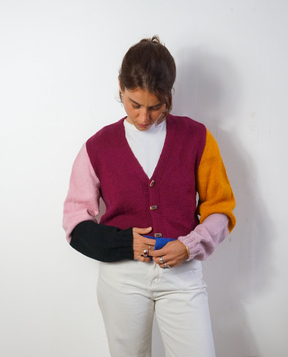 A woman wearing a color-blocked knit cardigan sweater with magenta, light pink, yellow, and black sleeves, paired with white pants, standing against a plain white wall.