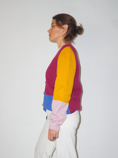 A woman shown in profile wearing a color-block knit cardigan with magenta, yellow, light pink, and blue panels, paired with white pants, against a plain white wall.