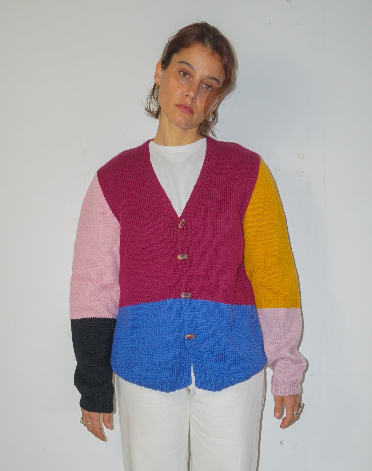 A woman wearing a color-blocked knit cardigan sweater with magenta, pink, yellow, and blue panels, styled over a white t-shirt and white pants against a plain white wall.