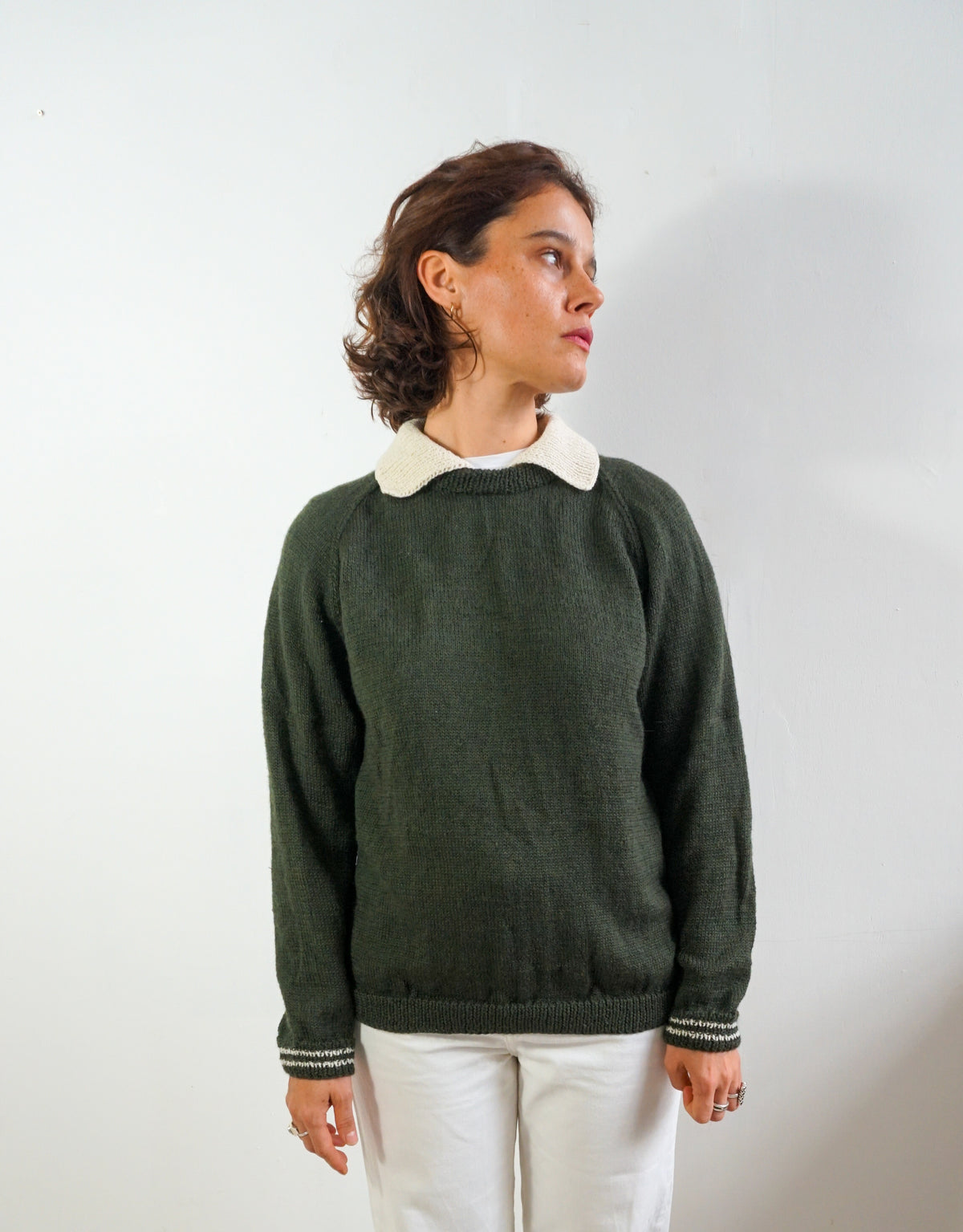 A woman wearing a dark green knit sweater with a contrasting cream-colored collar, paired with white pants, standing against a plain light gray wall.