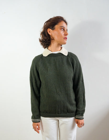 A woman wearing a dark green knit sweater with a contrasting cream-colored collar, paired with white pants, standing against a plain light gray wall.
