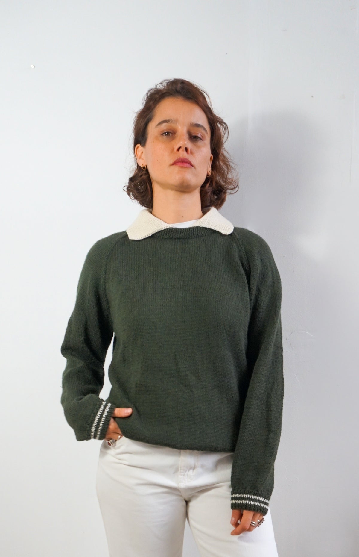 A woman wearing a dark green knit sweater with a contrasting cream collar and striped ribbed cuffs, paired with white pants, standing against a plain light gray wall.