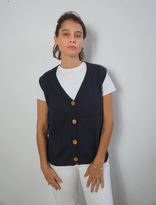 A woman wearing a dark navy blue knit button-up vest over a white t-shirt and white pants, standing against a plain light gray wall.