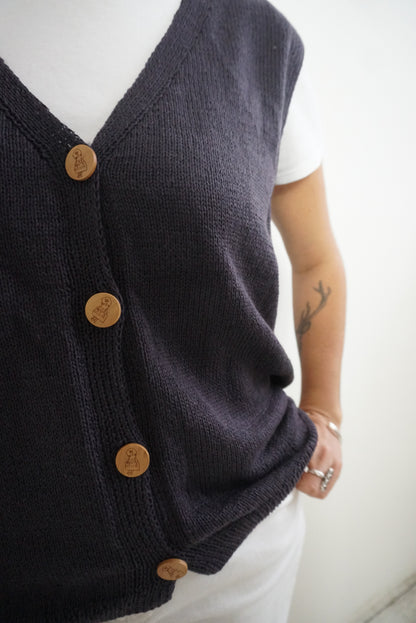 A close-up of a person wearing a dark charcoal knit cardigan vest over a white t-shirt. The cardigan features light brown wooden buttons down the front.