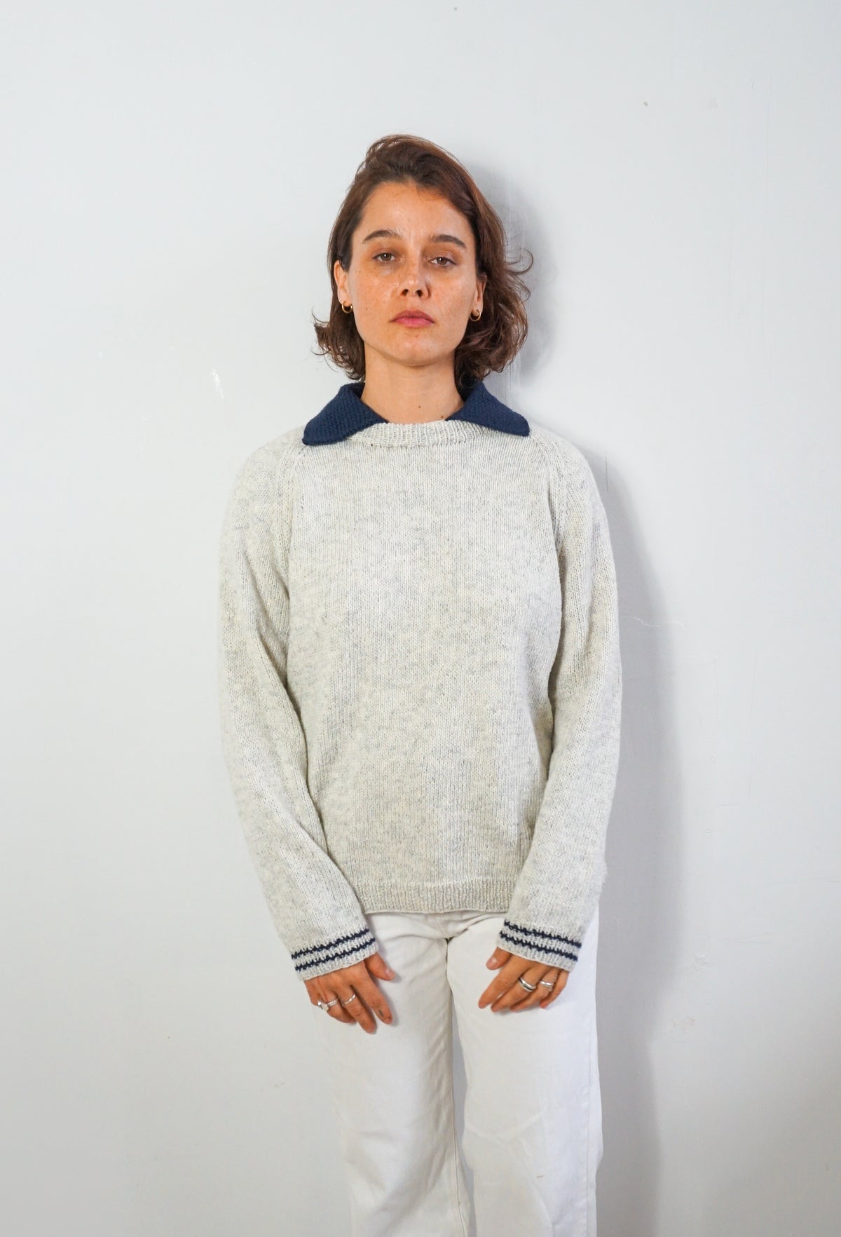 A woman wearing a light gray knit sweater with a contrasting dark navy blue collar and matching navy striped cuffs, paired with white pants, standing against a plain light gray wall.
