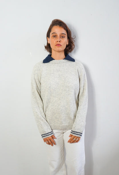 A woman wearing a light gray knit sweater with a contrasting dark navy blue collar and matching navy striped cuffs, paired with white pants, standing against a plain light gray wall.