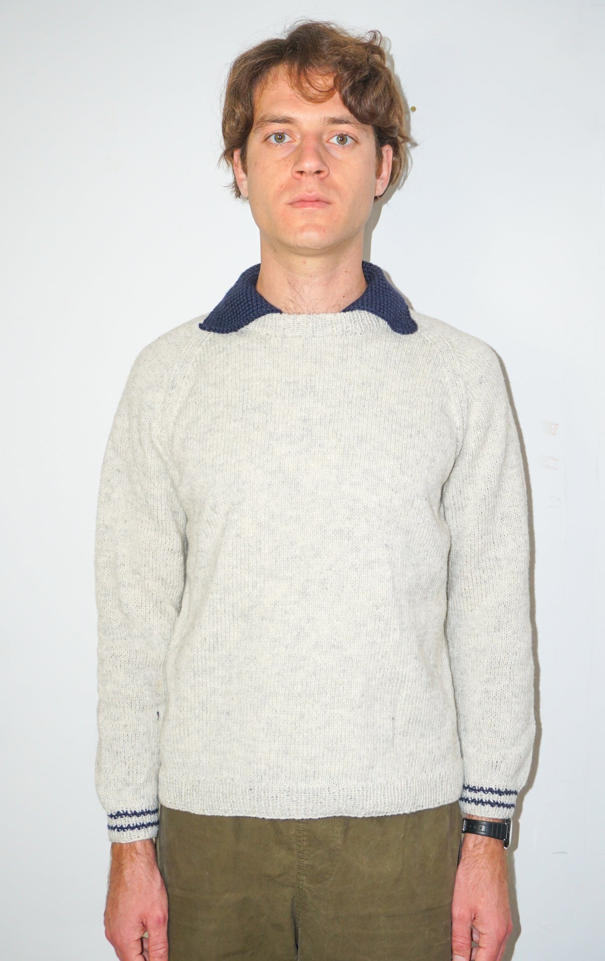 A man wearing a light beige knit sweater with a contrasting dark navy collar and matching navy striped cuffs, paired with olive green pants, standing against a plain white background.