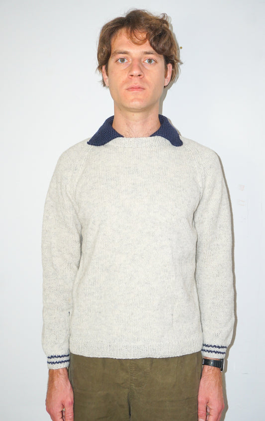 A man wearing a light beige knit sweater with a contrasting dark navy collar and matching navy striped cuffs, paired with olive green pants, standing against a plain white background.