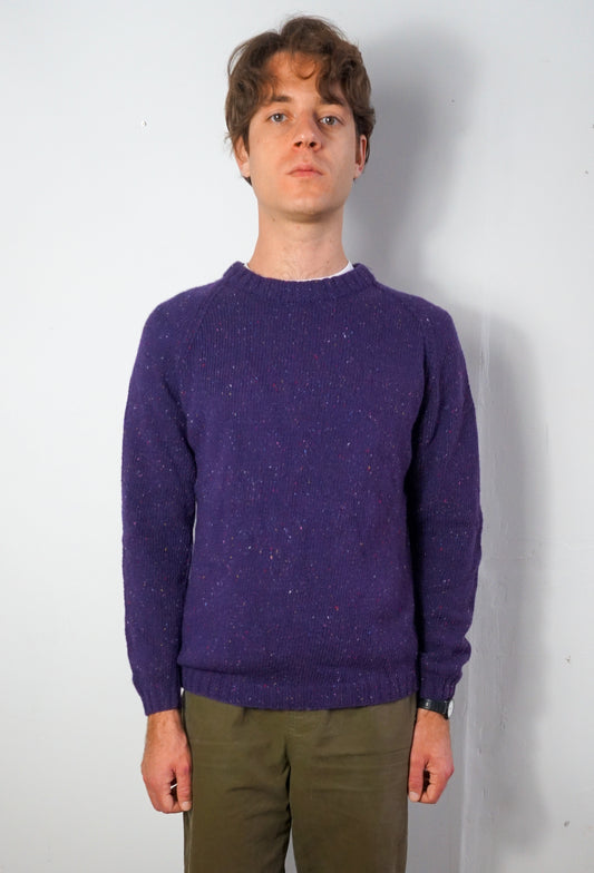 A man wearing a textured purple crewneck sweater and olive green pants, standing against a plain light gray wall.