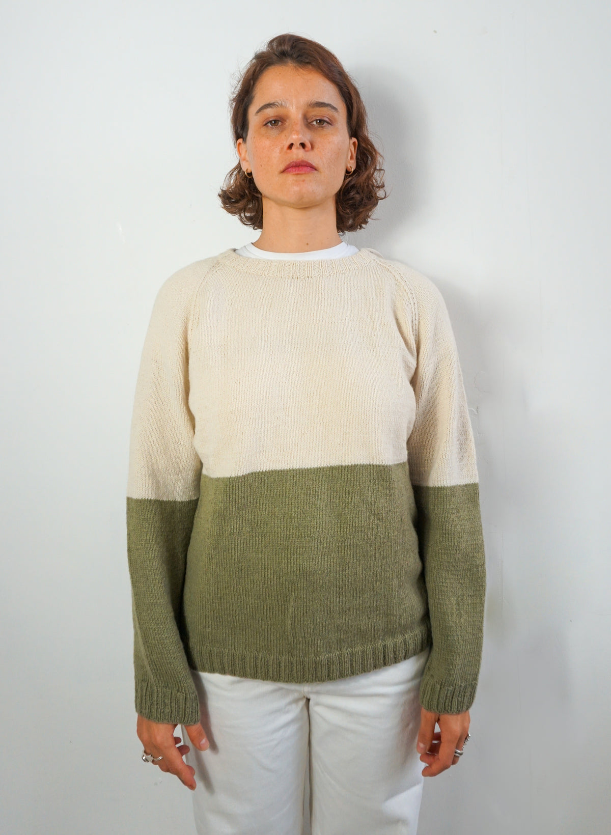 A woman wearing a colorblock knit sweater with a cream upper section and an olive green lower section and sleeves, paired with white pants, standing against a plain light gray wall.