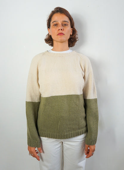A woman wearing a colorblock knit sweater with a cream upper section and an olive green lower section and sleeves, paired with white pants, standing against a plain light gray wall.