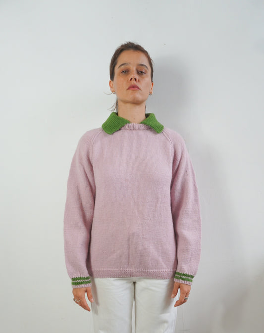 A woman wearing a light pink knit sweater with a contrasting green collar and green striped cuffs, paired with white pants, standing against a plain light gray wall.