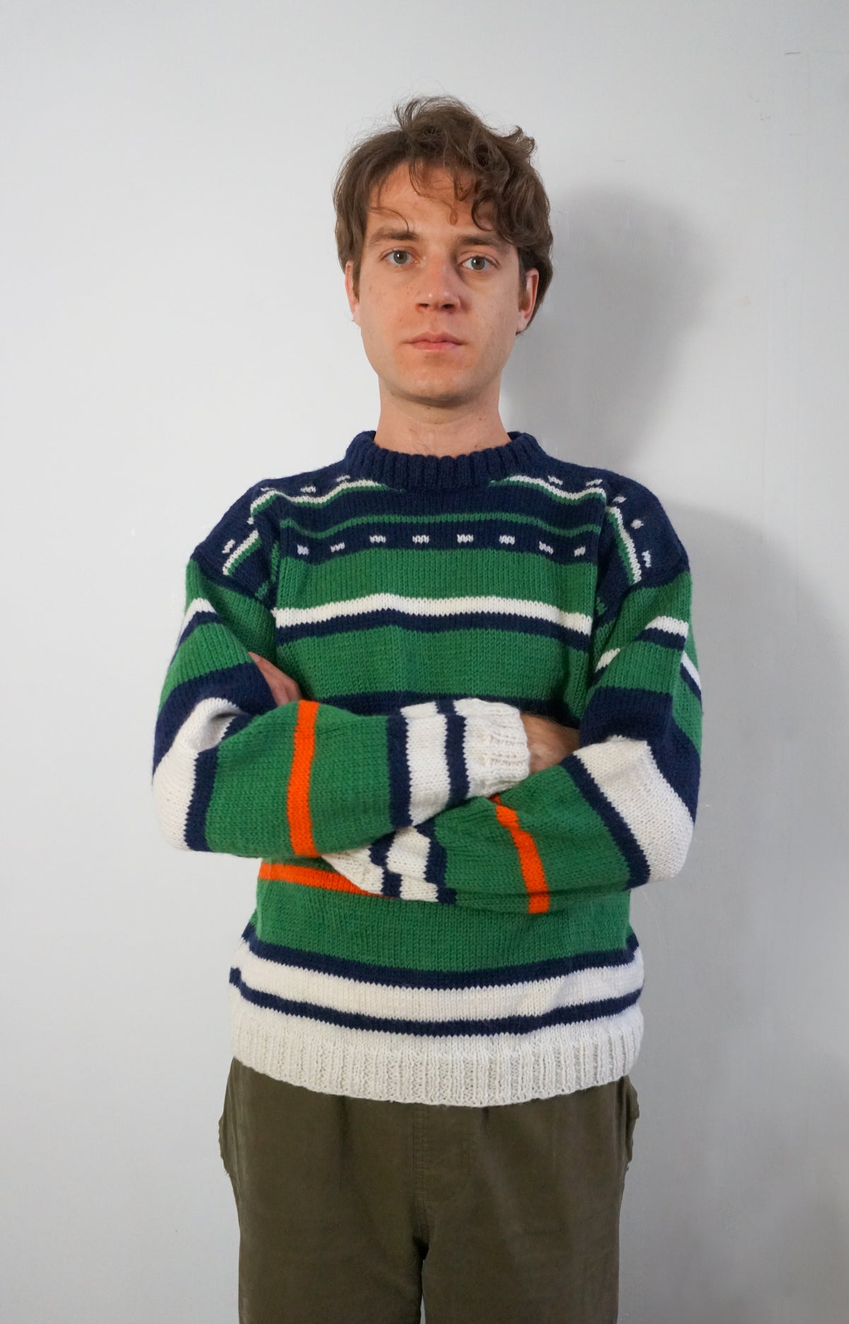 A man wearing a green, navy, and white striped knit sweater with orange accents, paired with olive green pants, standing against a plain light gray wall.