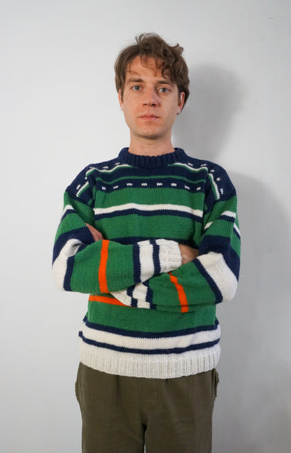 A man wearing a green, navy, and white striped knit sweater with orange accents, paired with olive green pants, standing against a plain light gray wall.