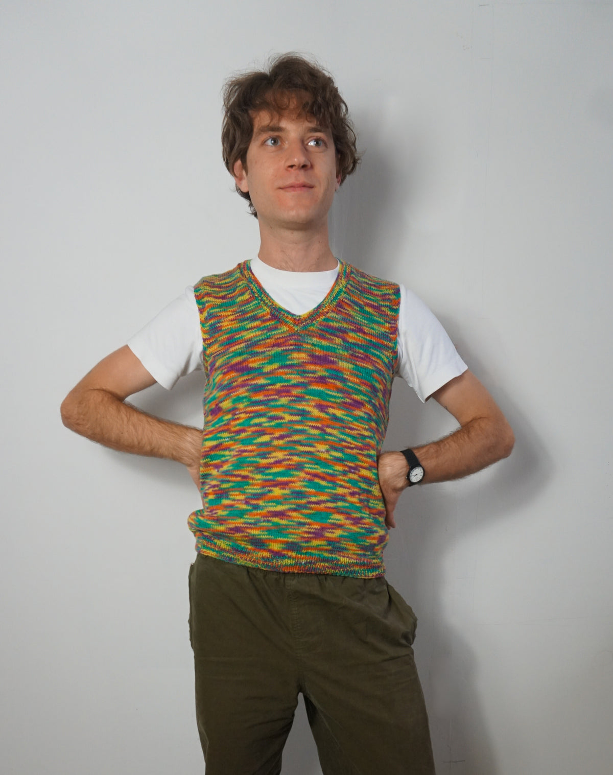 A man wearing a colorful, multicolor knit V-neck sweater vest over a white t-shirt and olive green pants, standing against a plain light gray wall.