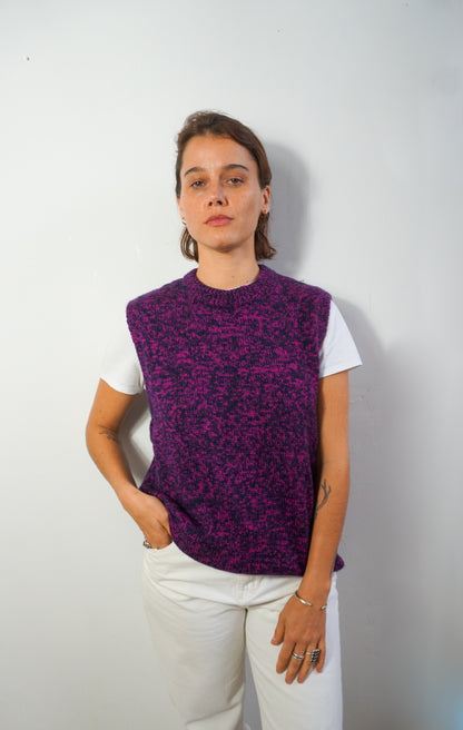 A woman wearing a purple and black marled knit sweater vest over a white t-shirt and white pants, standing against a plain light gray wall.