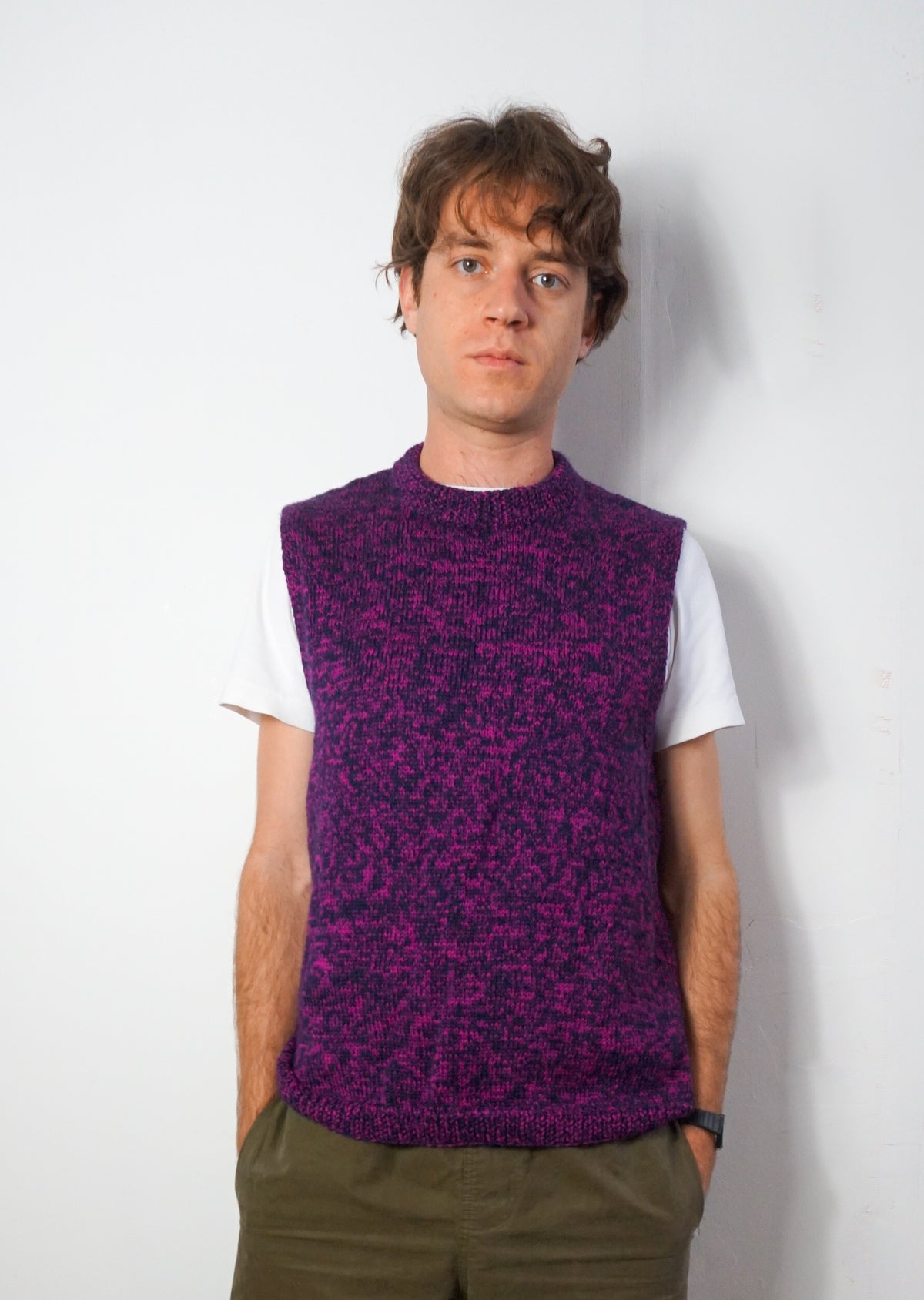 A man wearing a purple and black marled knit sweater vest over a white t-shirt and olive green pants, standing against a plain white wall.
