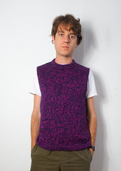 A man wearing a purple and black marled knit sweater vest over a white t-shirt and olive green pants, standing against a plain white wall.