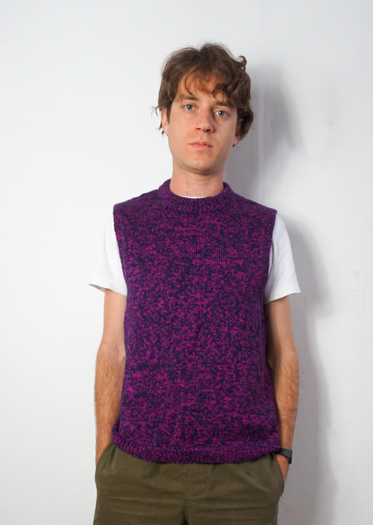 A man wearing a purple and black marled knit sweater vest over a white t-shirt and olive green pants, standing against a plain white wall.