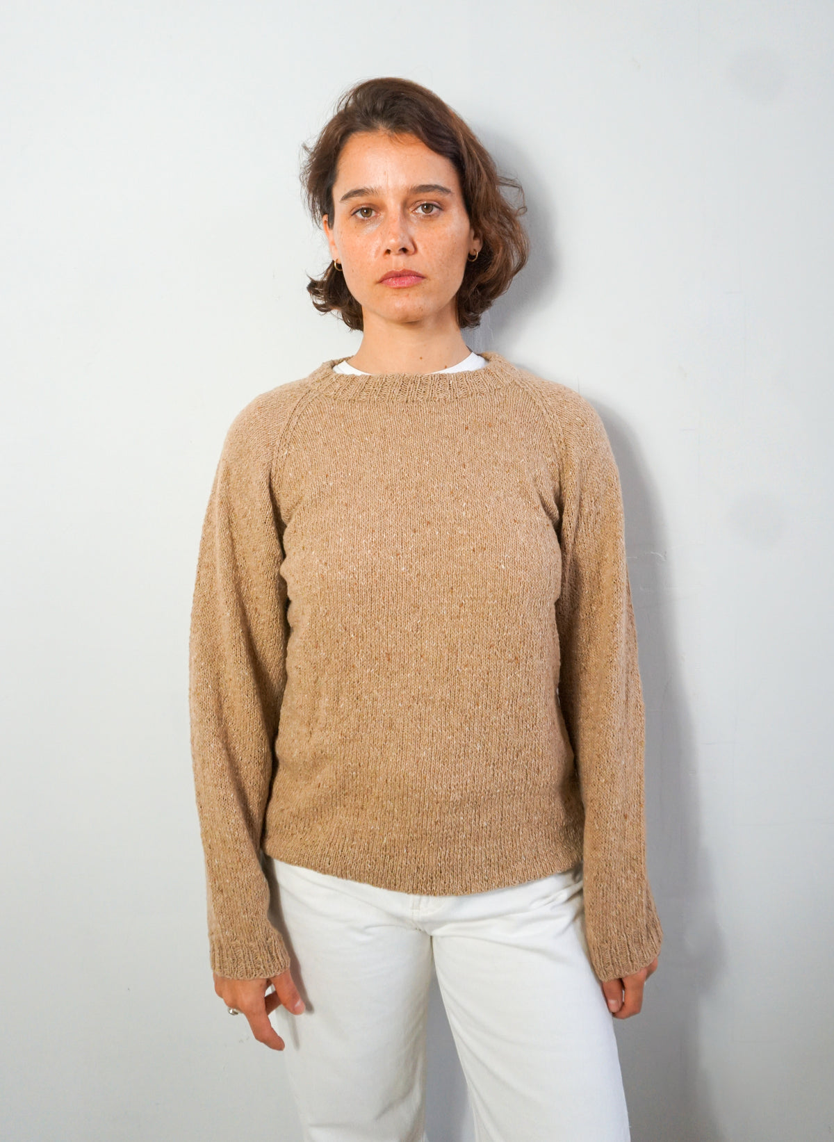 A woman wearing a beige knit crewneck sweater and white pants, standing against a light gray wall.