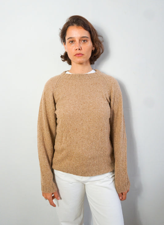 A woman wearing a beige knit crewneck sweater and white pants, standing against a light gray wall.