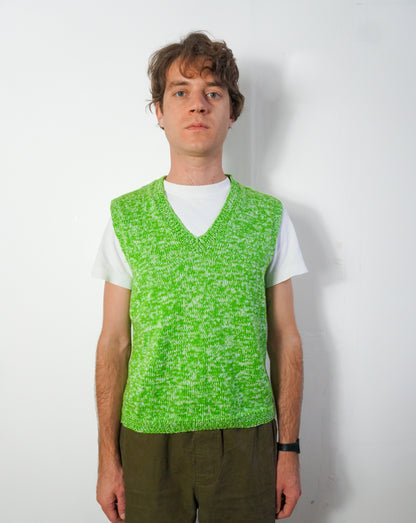 A man wearing a bright green and white marled knit sweater vest over a white t-shirt and olive green pants, standing against a plain white wall.