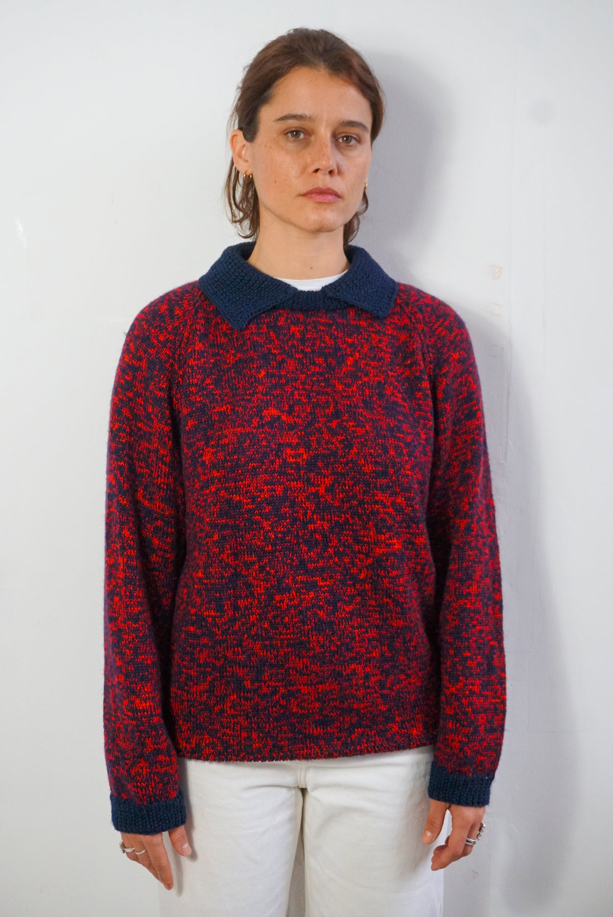 A woman modeling a vintage knit sweater with a red and navy blue mottled pattern, featuring a solid navy blue collar and ribbed cuffs, paired with white pants against a plain white wall.