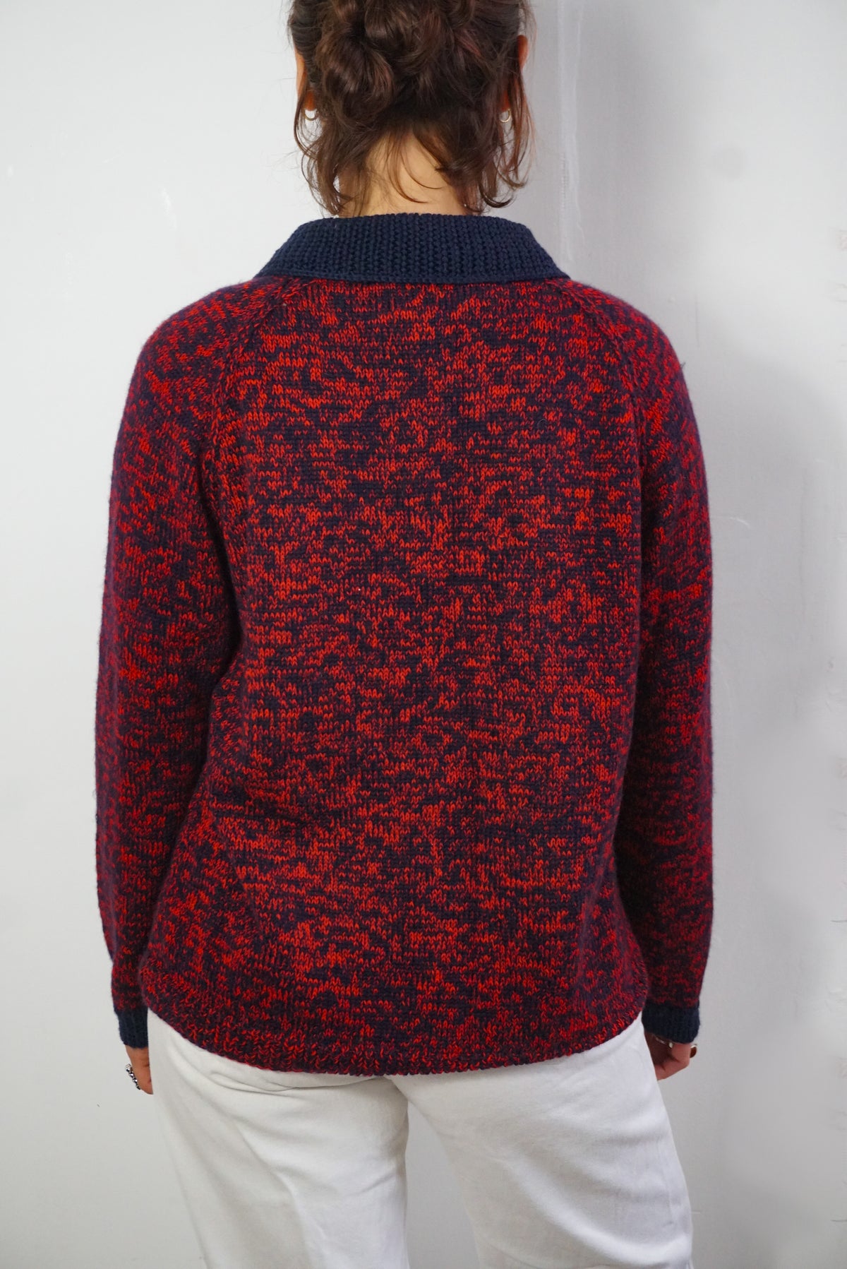 A person viewed from the back wearing a red and navy marled knit sweater with a solid navy ribbed collar, paired with white pants, against a plain white wall.