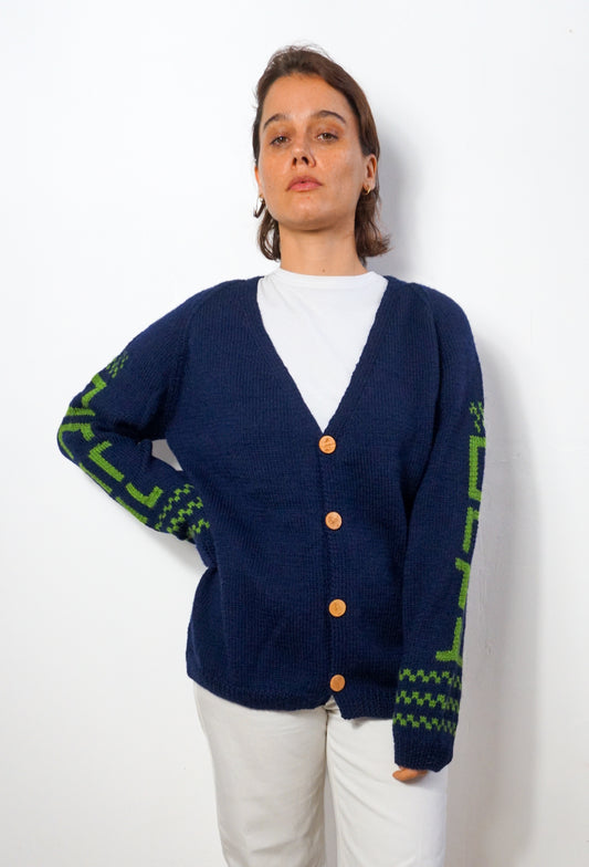 A woman wearing a navy blue knit cardigan with green geometric patterns on the sleeves and hem, styled over a white t-shirt and white pants, standing against a plain white wall.