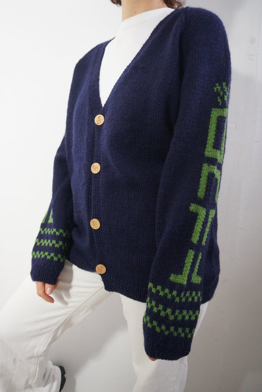 A person wearing a navy blue knit cardigan with green geometric patterns on the sleeves and cuffs, paired with a white top and white pants, photographed against a plain light background.